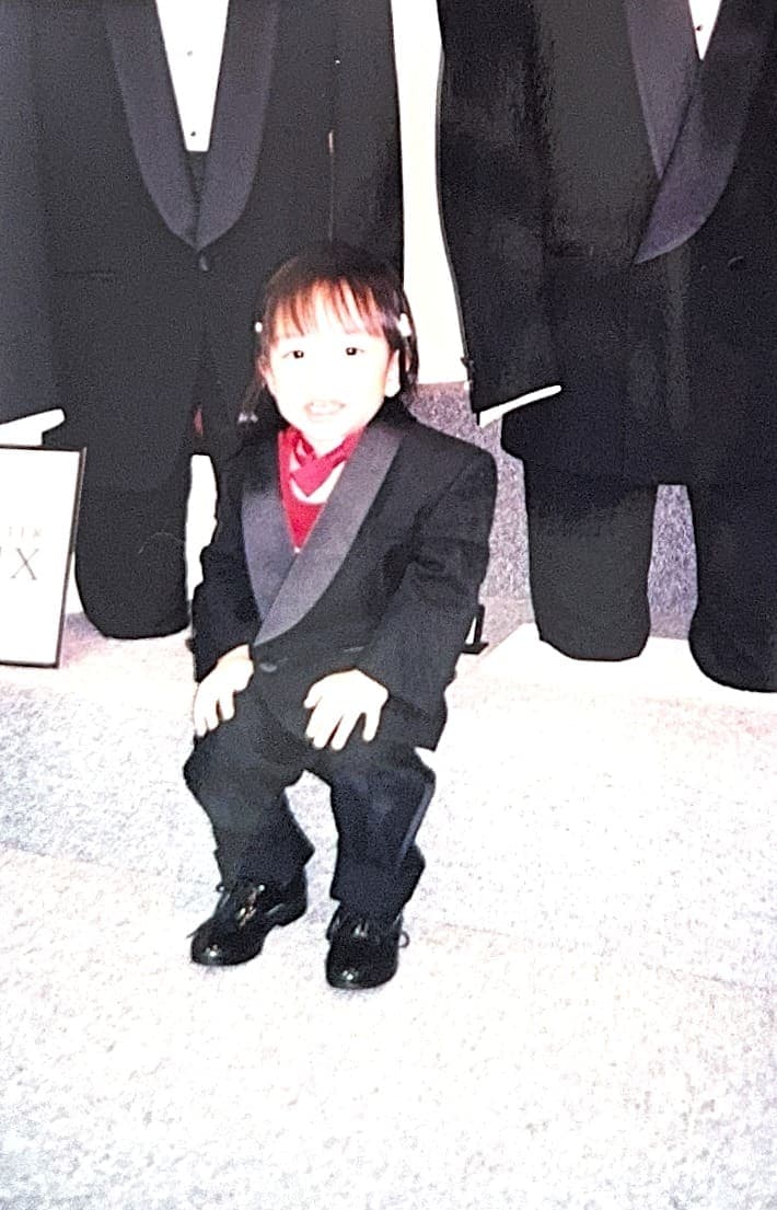 Young Lauren in a child's tuxedo among the hanging suits